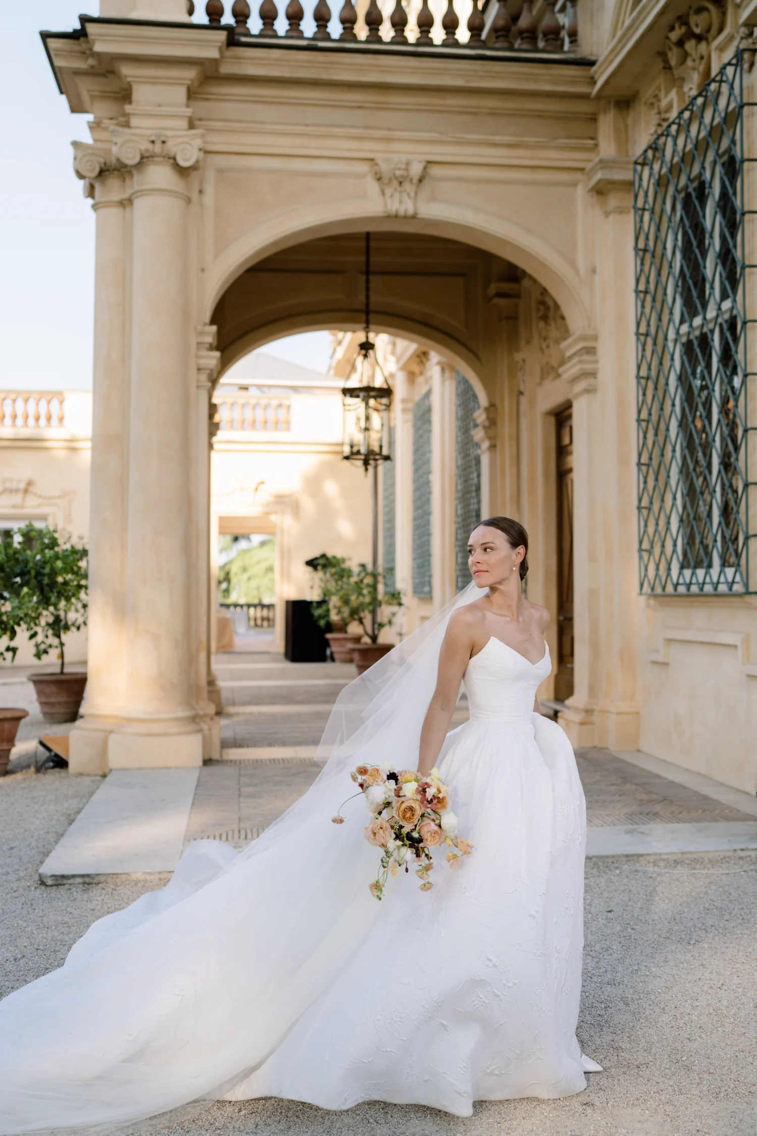 Bride under a stone archway holding a loose bouquet in cream and apricot